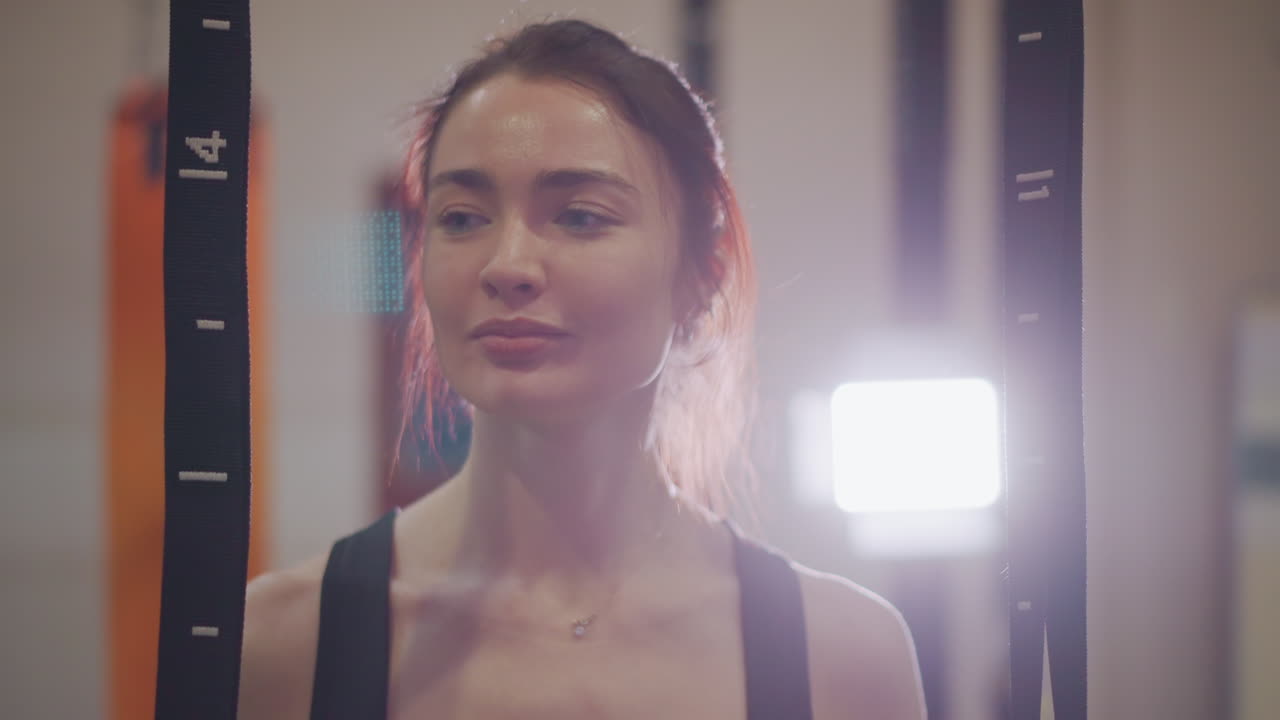 Ritratto in palestra di donna sorridente sotto le luci, aureola di capelli retroilluminata, bagliore morbido dell'obiettivo, defaticamento post-allenamento, gioia e sollievo sottili, spalle rilassate, movimento di stretching, atmosfera da studio con allenatore.