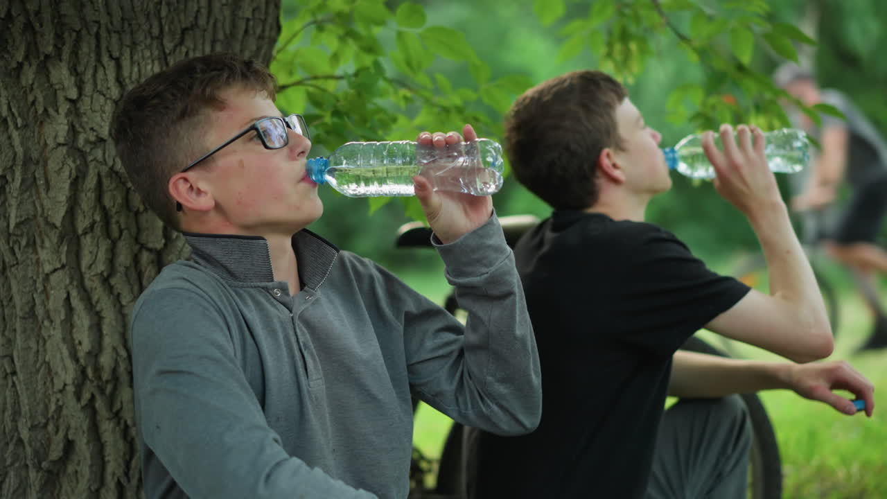 두 어린 형제는 나무 에 앉아서 자전거 타고 휴식을 취하는 동안 물병에서 물을 마시고 자전거를 타고 지나가는 누군가의 흐릿한 시야와 함께 근처에 주차되어 있습니다.