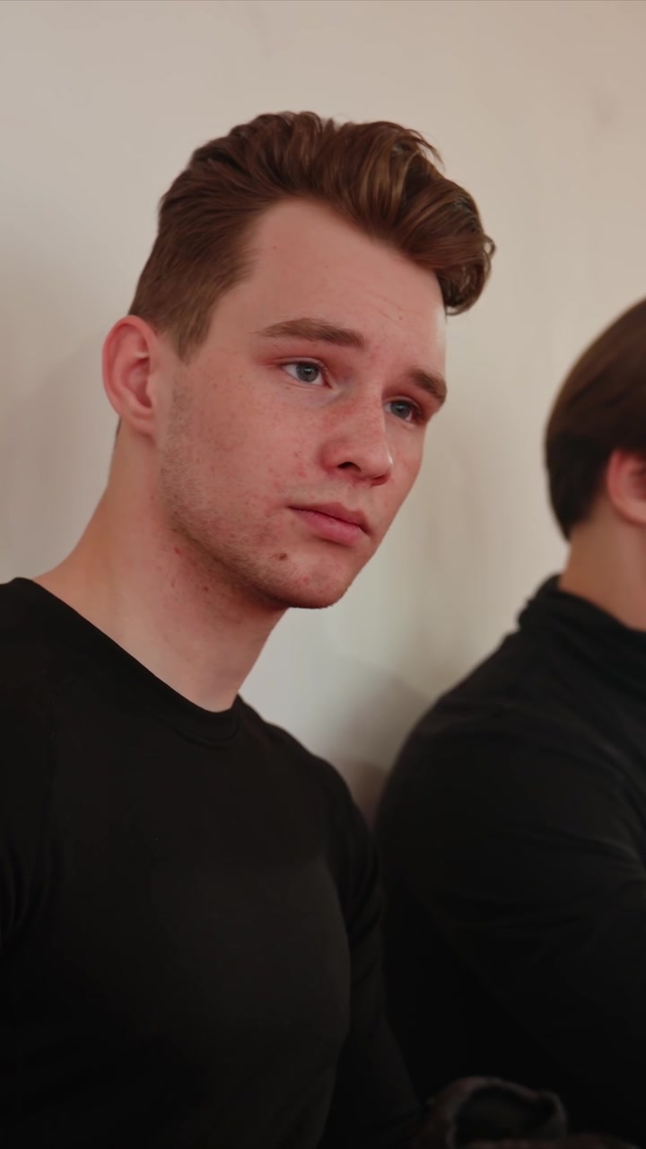 Primo piano di un giovane uomo con capelli castani che indossa una camicia nera. L'uomo gira leggermente la testa mentre è seduto, in modo discreto. Entrambe le persone rimangono per lo più immobili durante l'intera sequenza ambientata al chiuso.
