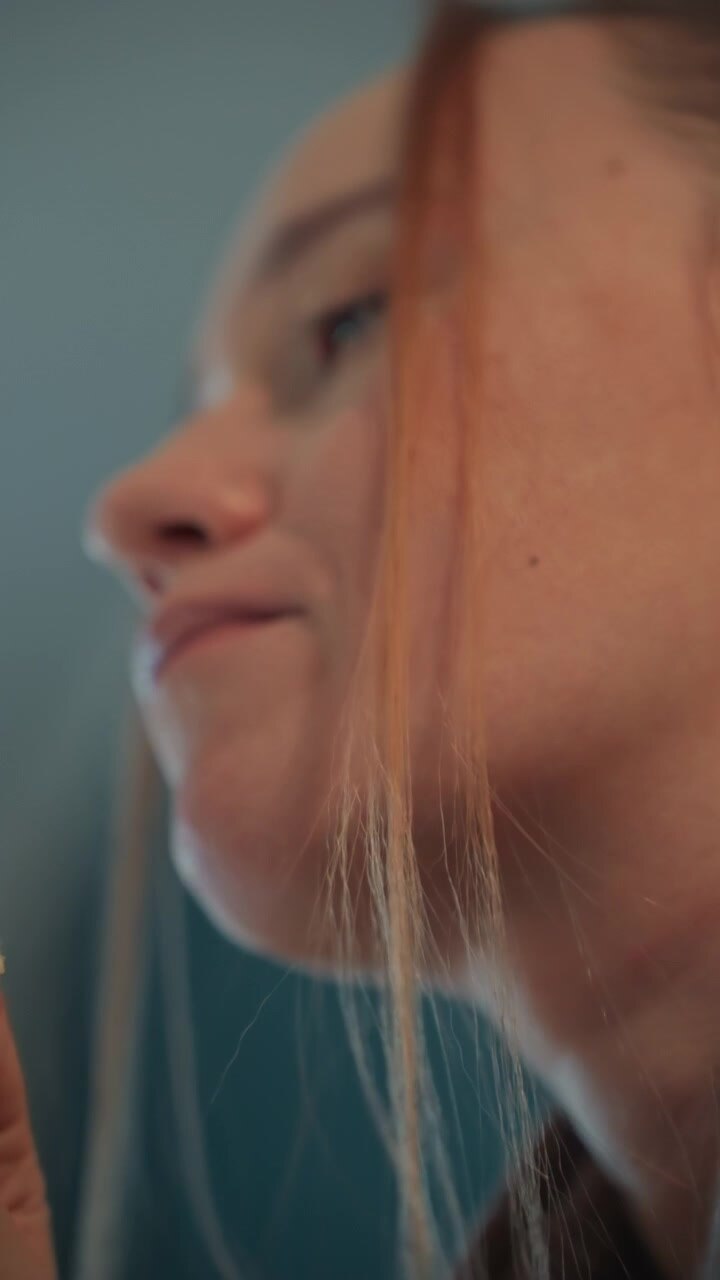 Close up of woman eating small food items focusing on their profile. Subject holds various snacks near mouth with minimal movement. Background is soft blue gray indoor setting