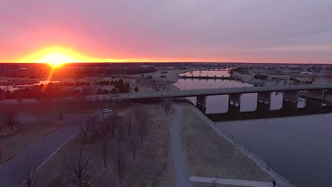 fly-over-of-trail-along-rivers-edge-towards-bridge-with-sunset-in-background