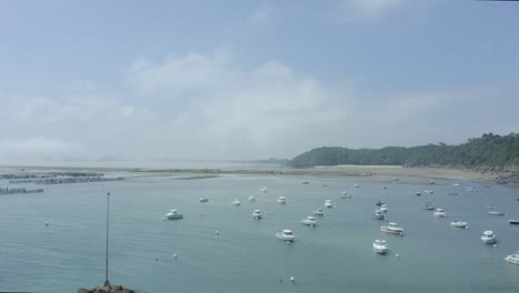 Vuelo-De-Drones-Desde-Barcos-Que-Flotan-En-La-Superficie-Del-Mar-Hasta-Una-Granja-De-Ostras-Con-Granjeros-Y-Tractores-Ocupados-Trabajando-En-Un-Día-Soleado-En-Bretaña,-Francia