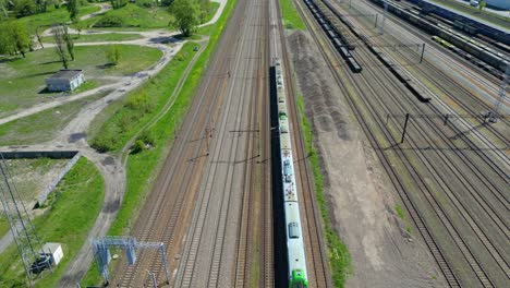 Toma-Aérea-Panorámica-De-Un-Tren-De-Pasajeros-Conduciendo-Hacia-El-Atardecer-En-Vías-De-Tren-Rodeadas-De-árboles-Sobre-El-Paisaje-Urbano-Hacia-Adelante