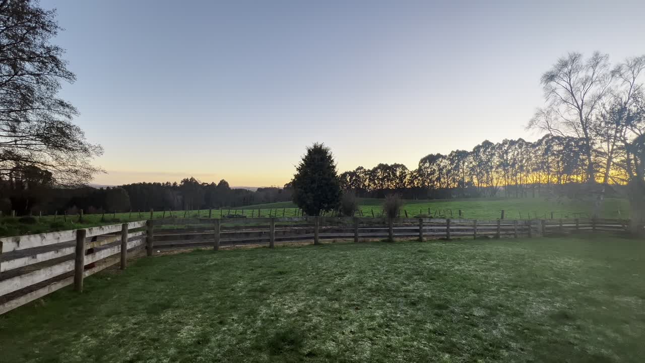 A serene New Zealand farm landscape at sunset, featuring a rustic wooden fence, lush green fields, and distant trees under a quiet, colorful sky. Perfect for relaxation and nature-themed content.