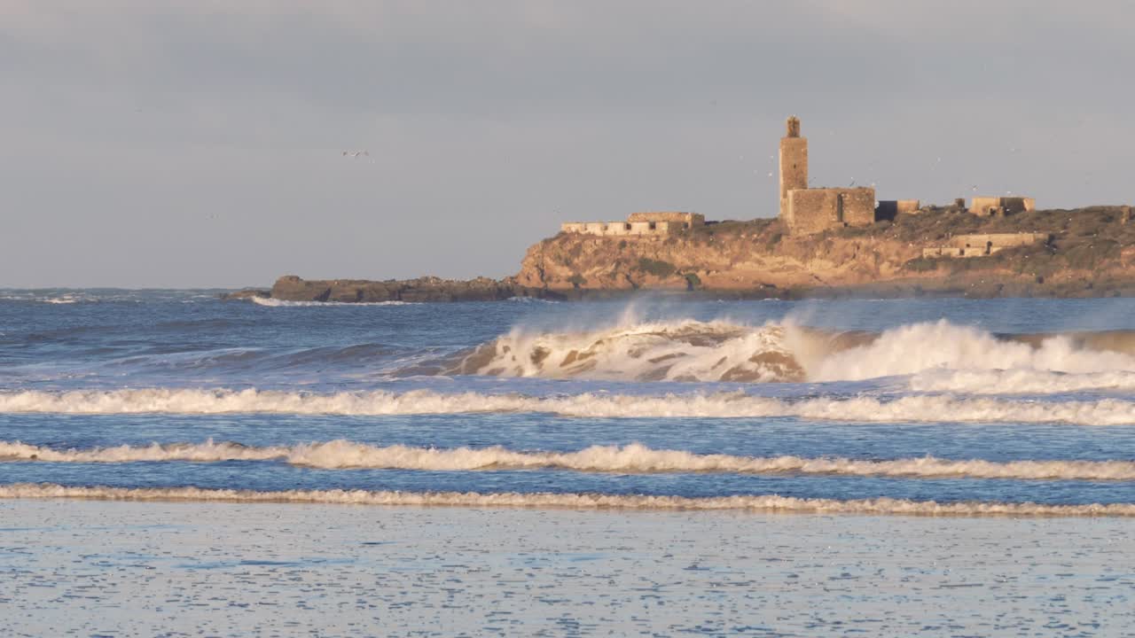 altas olas marinas en la playa de essaouira en marruecos con vista a una isla con mezquita y edificios antiguos, ruinas.