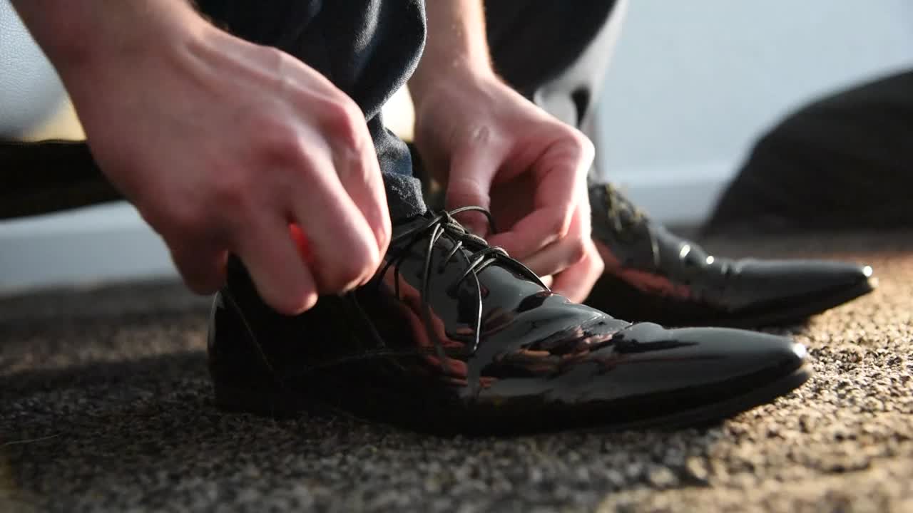 Side view of man tying laces on black, shiny dress shoes on dark carpet. Inside