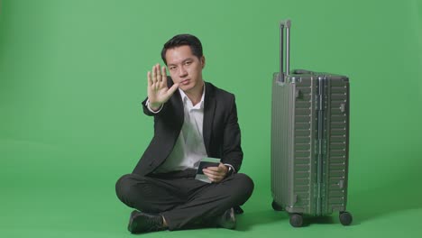 full body of asian businessman traveler with luggage and passport looking at camera and disapproving with no hand sign in the green screen background studio