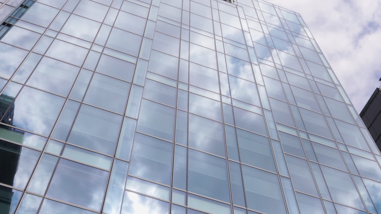 The towering glass facade of a modern building in Canary Wharf reflects the dynamic sky, creating a sleek architectural view. Clear daylight enhances the urban atmosphere