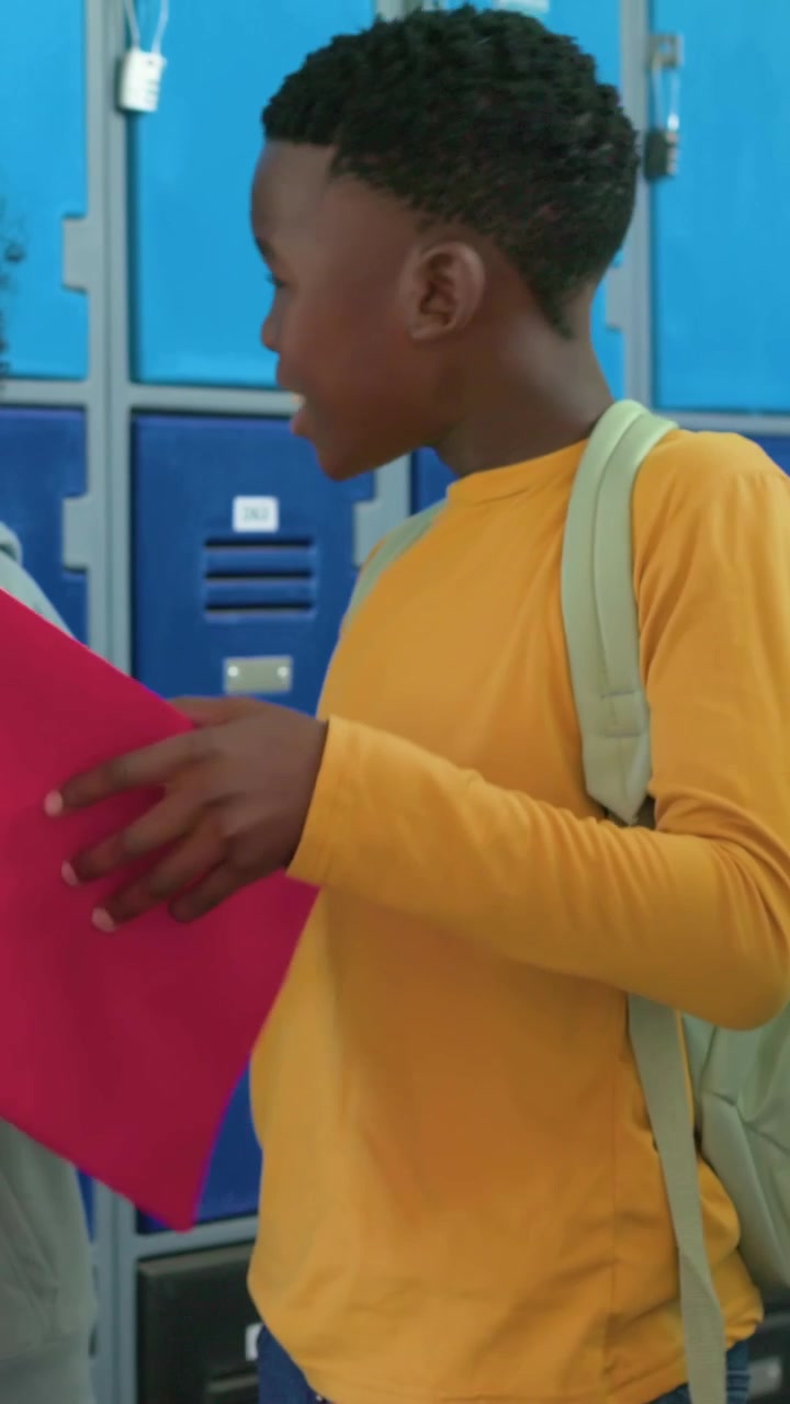 Vertical video: Receiving pink folder from peer, student in mustard shirt checking pages at lockers