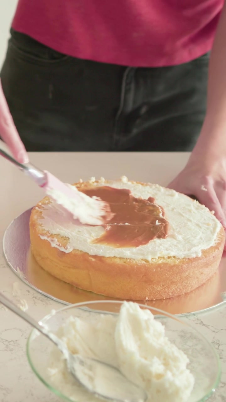 Video verticale: Mette una torta sul bancone, lei glassa e spalma il cioccolato per decorare, con un top rosa.