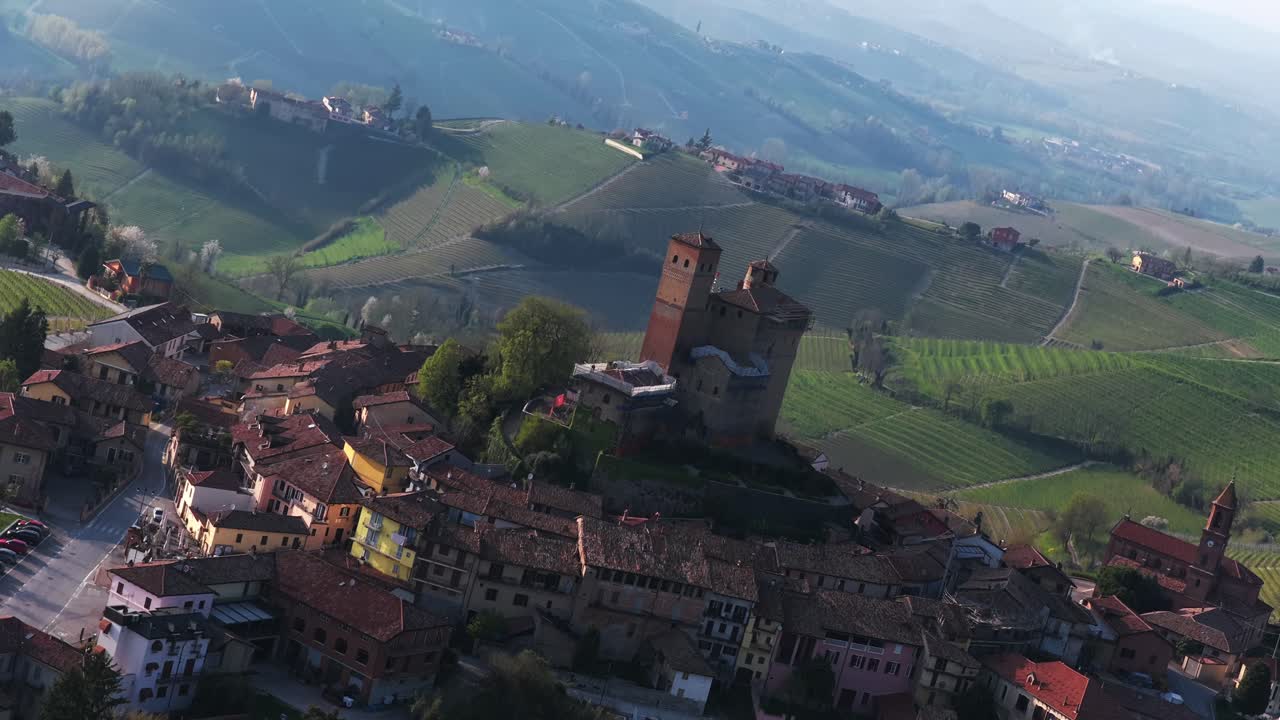 Dutch angle aerial shot of Serralunga d'Alba, emphasizing the medieval castle at the center, surrounded by tightly packed village roofs and vibrant vineyards stretching across the rolling hills