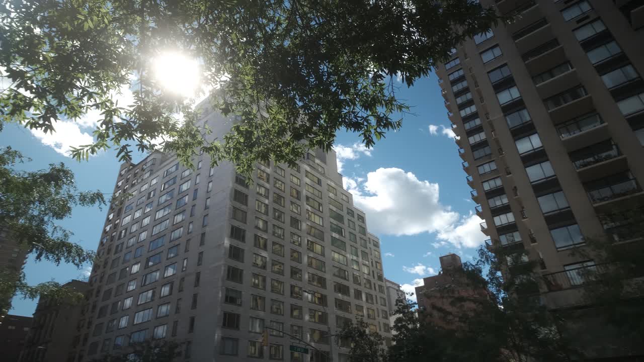 clear upward pan of tall apartment buildings in New York City revealing the sun and a lens flare through trees