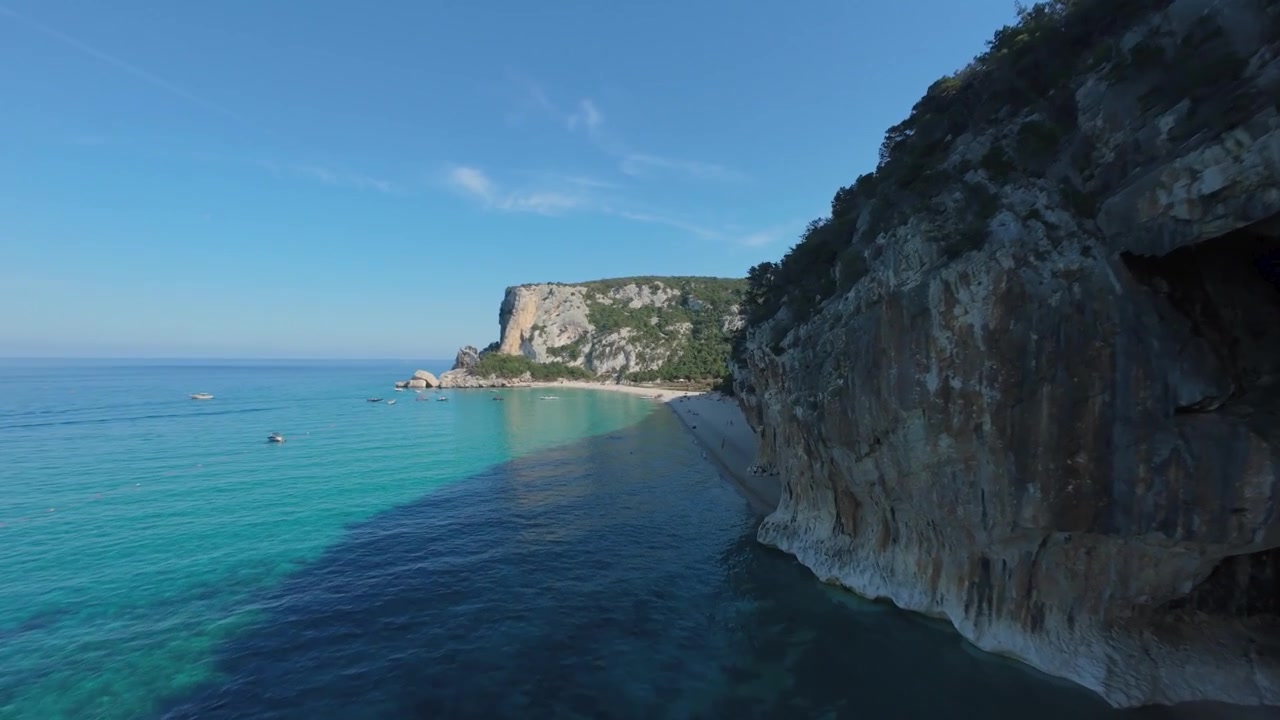 FPV-drone lento matalalla turkoosin veden yllä Cala Lunan kalkkikivikallioiden vierellä, Baunein rannikolla, Sardiniassa. Meriluolat, kristallinkirkas Välimeri, dramaattinen rannikko, kesäkuun kesävalo