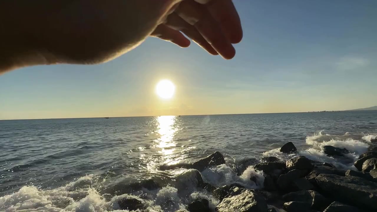 Hand Blocking Golden Sun Over Ocean Waves and Rocks