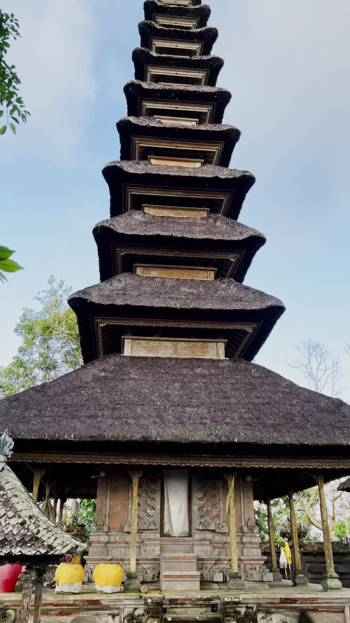 Vertikalt billede af traditionel balinesisk hindu tempelarkitektur med udskårne stendetaljer, ceremonielt alter og stråtag, beliggende i en fredfyldt gårdsplads omgivet af frodigt grønt på Bali