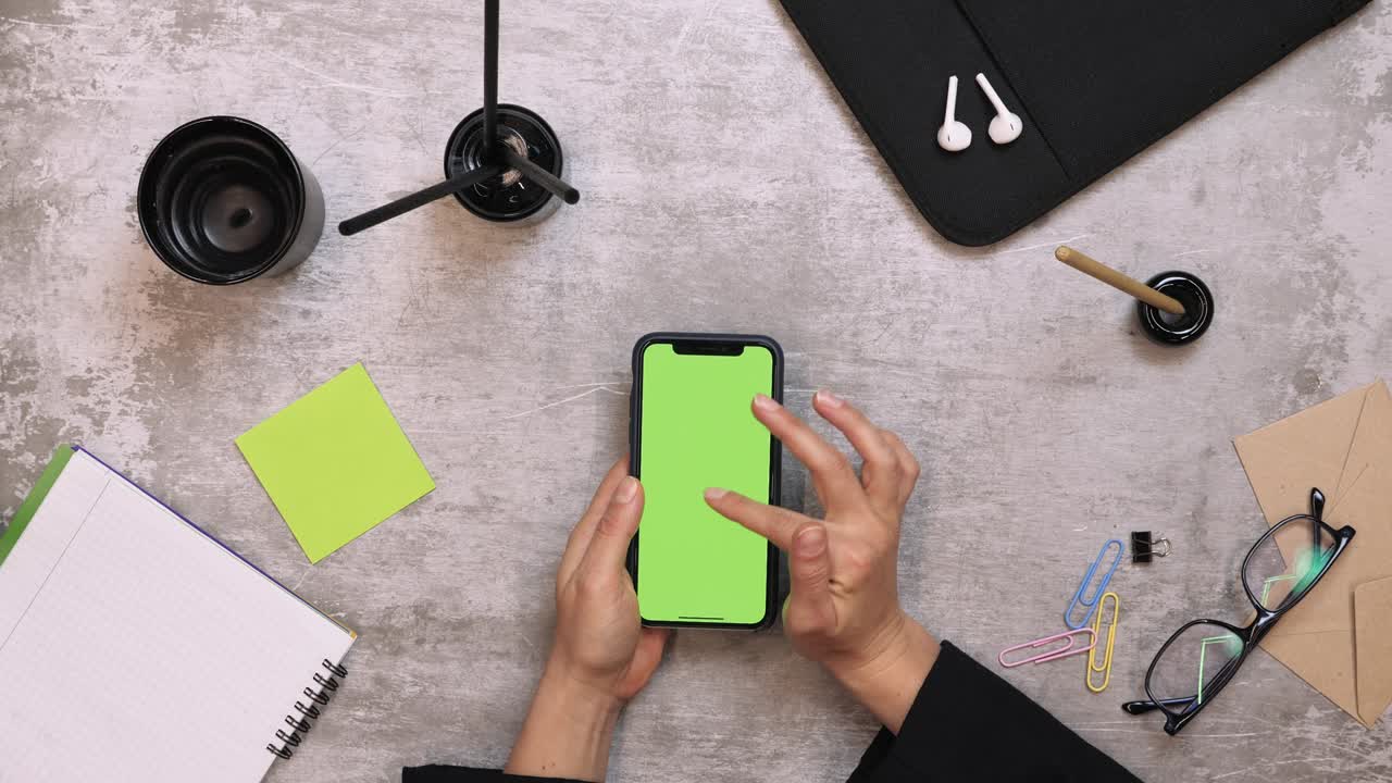 Woman using smartphone with green screen, scrolling news, tapping on photos, watching news in internet. Office desk background. Top view of office work desk. Female watching photos on smartphone