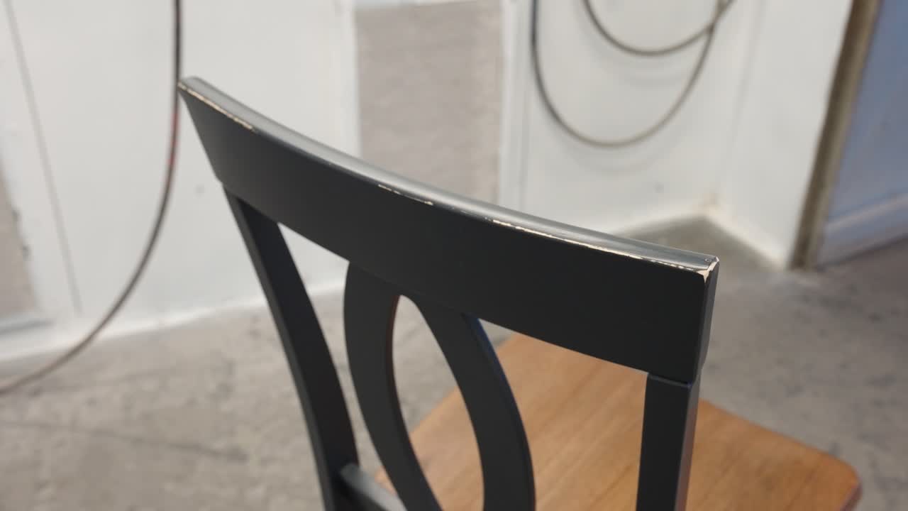 Still frame shows back of wooden dining chair with worn finish and bare wood