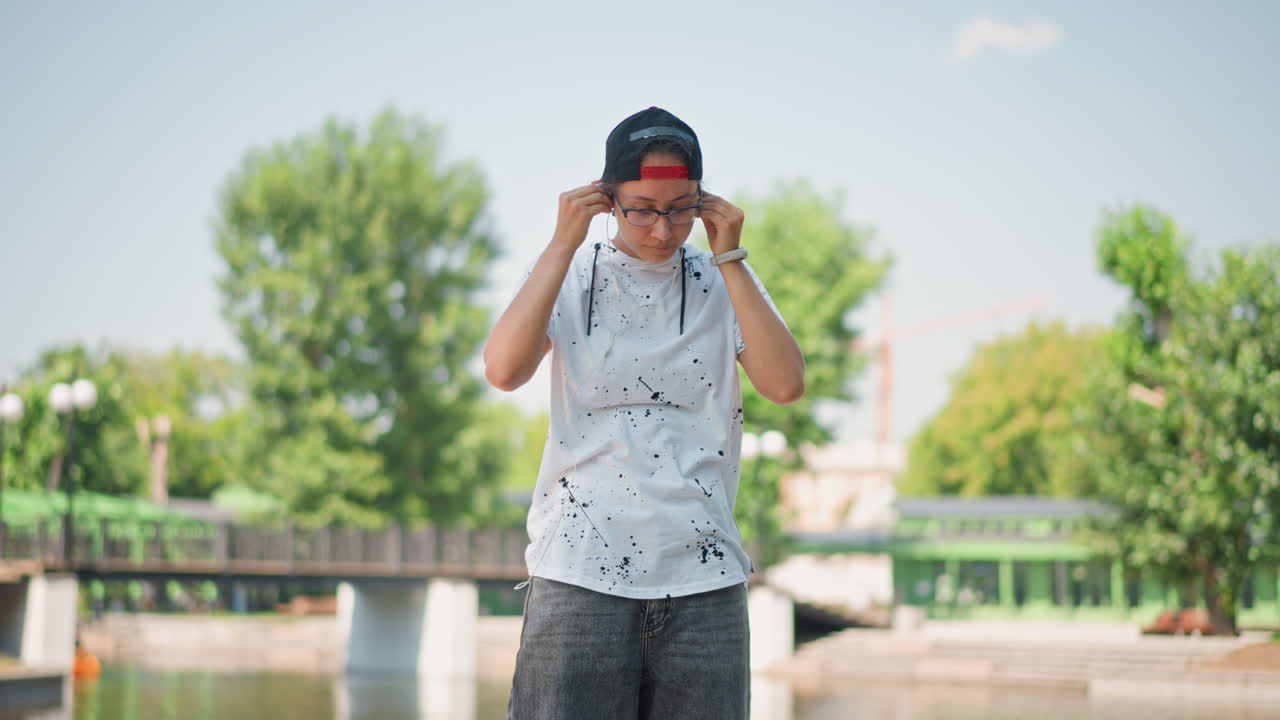 Teenage Asian male adjusting shades outdoors, Young Asian man confidently posing near river under sunlight, Casual young Asian male in playful pose adjusting sunglasses outside by water