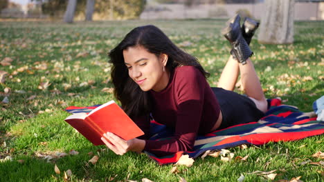 a young woman reading a book in the park during the autumn season with fall leaves blowing in the wind as she smiles