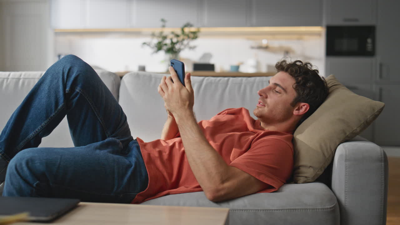 Stretching guy resting sofa at home. Joyful hipster taking mobile phone laying