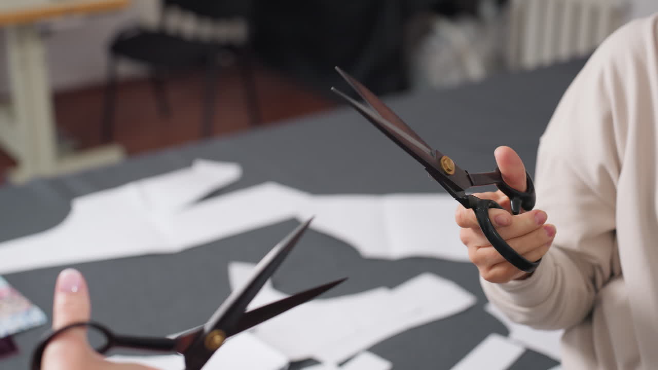 Dress makers hands holding sewing clipper engaging in playful scissors cutting fight above work table covered with sewing patterns and colorful fabric samples in bright creative workspace