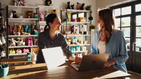 two women working and interacting at a shop counter with laptops