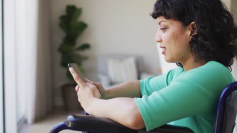 Happy-biracial-disabled-woman-in-wheelchair-using-smartphone-in-living-room