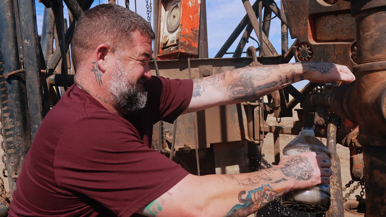 Man in purple t-shirt stands at the rusty pipes filling the bottle with dirty water. Site for drilling natural resources.