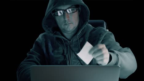 hacker in a hoody sweatshirt and wearing glasses is sitting at table at night and coding. hacker man removes memo from laptop screen. alpha channel chroma key transparent background.