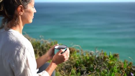 a serene moment of reflection by the ocean: a woman enjoys a cup of tea while gazing at the calm waves and beautiful landscape in tranquil surroundings
