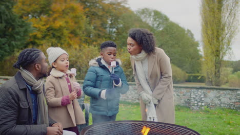 family blowing on toasted marshmallows to cool them sitting around firepit or barbeque in garden at home - shot in slow motion