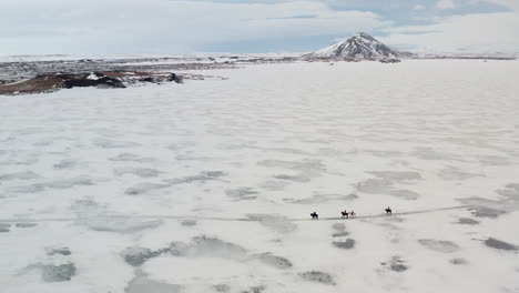 aerial winter landscape, group of tourists horses riding tour, trip on vast frozen lake myvatn in winter following path on ice in north iceland, vindbelgjarfjall mountain in horizon