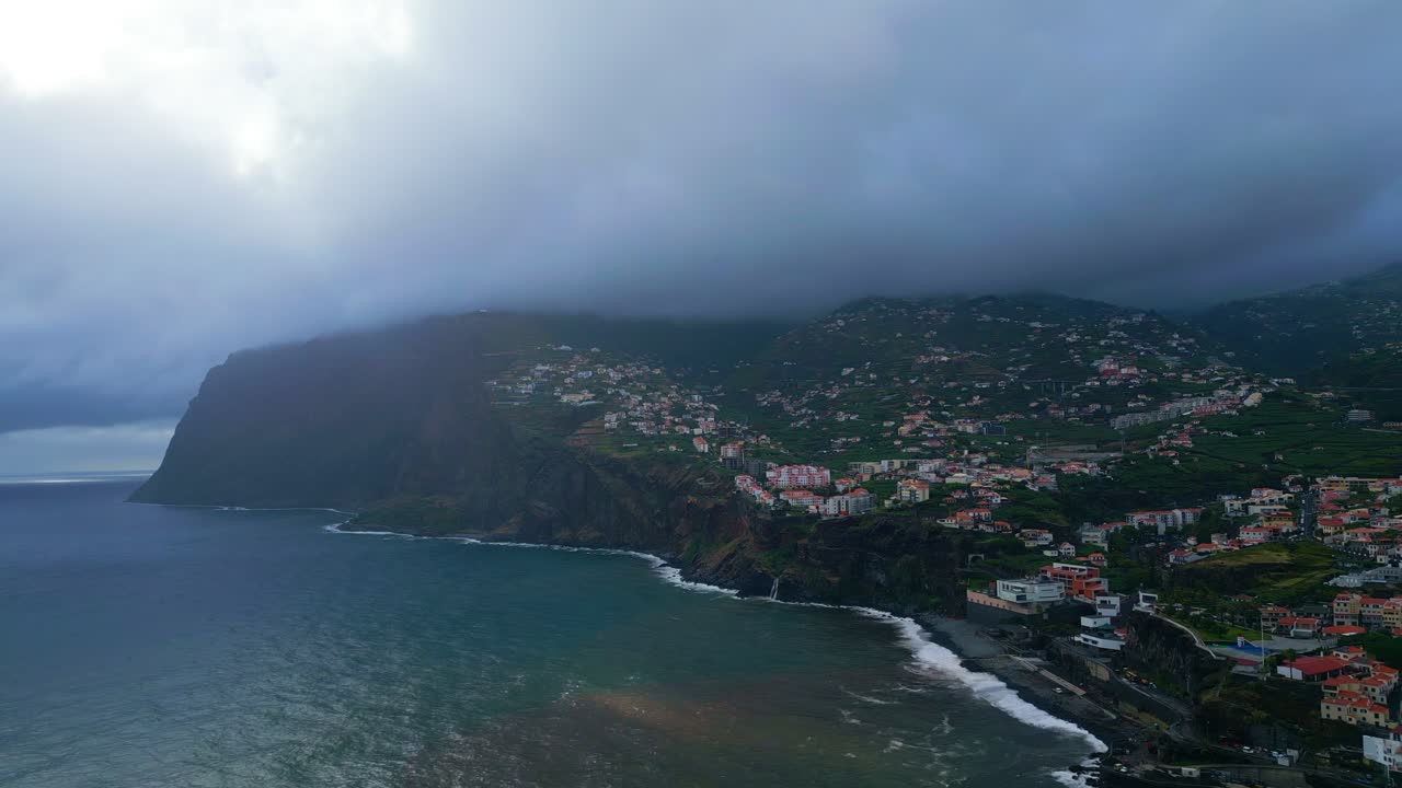 Filmato aereo di nuvole basse e nebbia che si estendono sulla costa a Praia Formosa a Funchal, Madeira. Scogliere suggestive, onde dell'Oceano Atlantico e quartieri costieri sotto un cielo plumbeo.