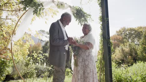 happy senior biracial couple exchanges rings during wedding ceremony in garden, slow motion