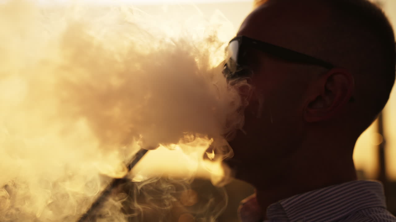 Silhouette of man in glasses smoking hookah sitting outdoors. Close up. Backdrop of setting sun.