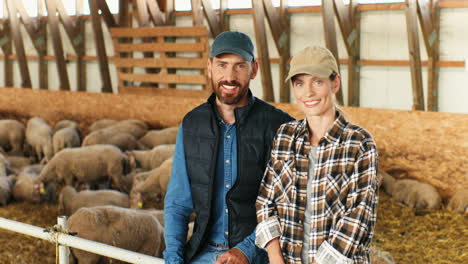 portrait of happy young caucasian married couple of farmers looking at each other and smiling at camera while sitting in stable with sheep flock