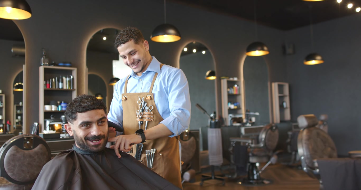 Frisør og kunde, mand i forklæde trimmer bakkenbarter med klippere i butikken, efter kunden har drejet hovedet.