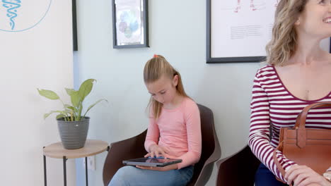 Caucasian-girl-patient-using-tablet-sitting-with-mother-talking-in-hospital-reception,-slow-motion