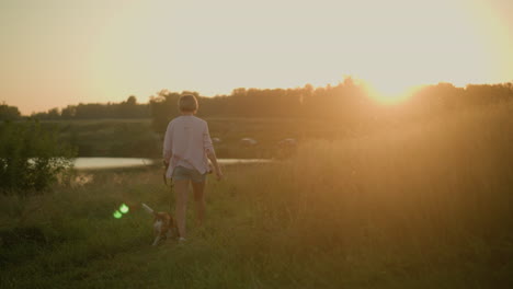 back view of woman walking her dog on leash during golden hour through grassy field, surrounded by natural scenery as sun sets in background, creating warm golden glow