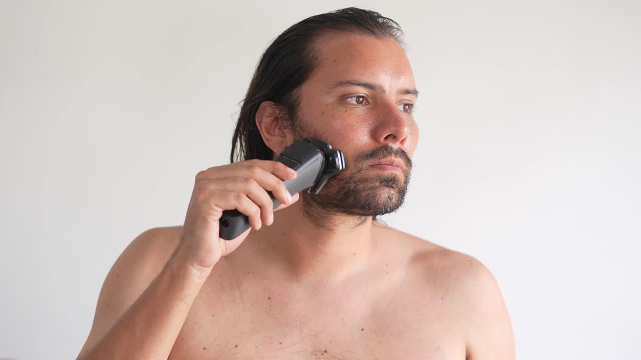 한 남자가 집에서 욕실에서 거울 앞에 서서 수염과 수염을 잘라내기 위해 전기 면도기를 사용하고 있습니다.