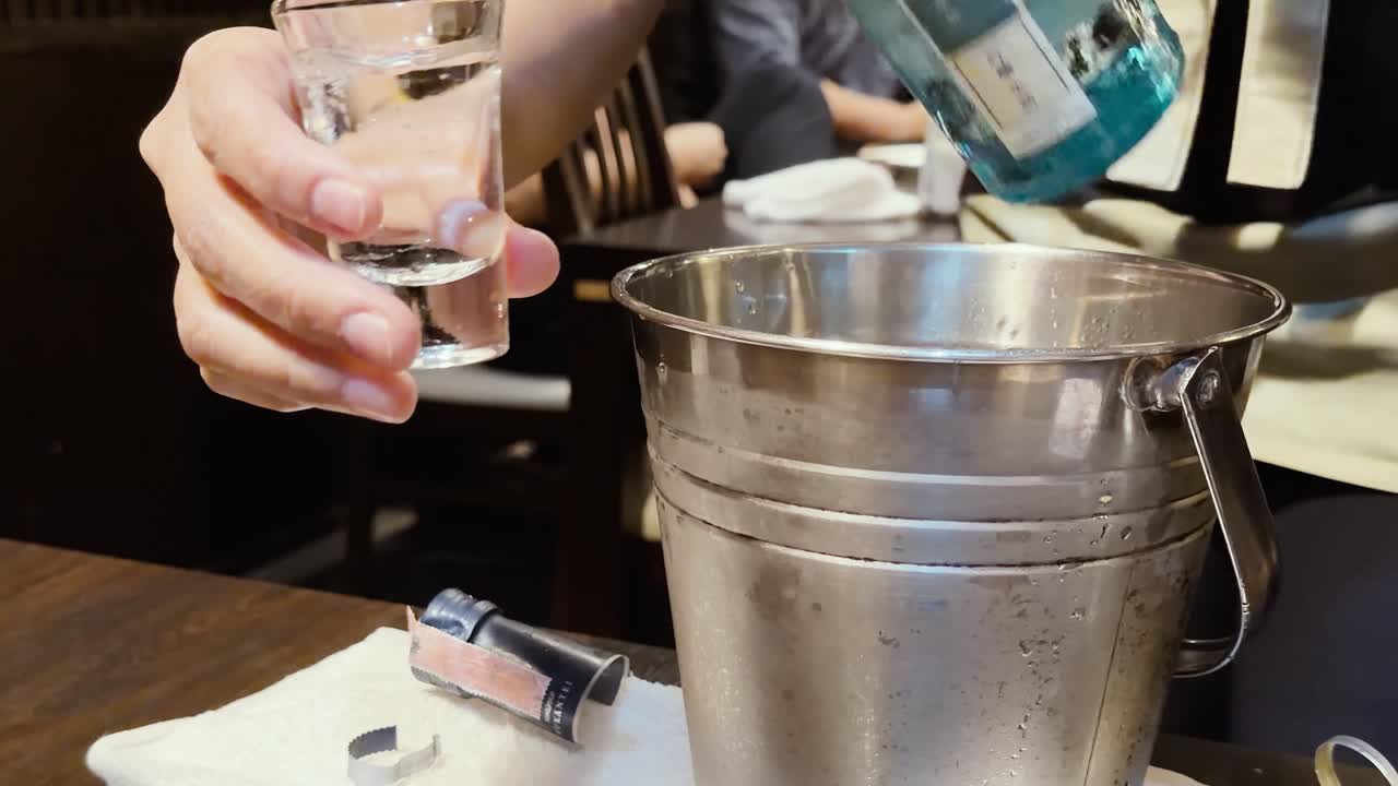 Primo piano di mani che versano sakè da una bottiglia blu in bicchieri accanto a un secchiello per il ghiaccio