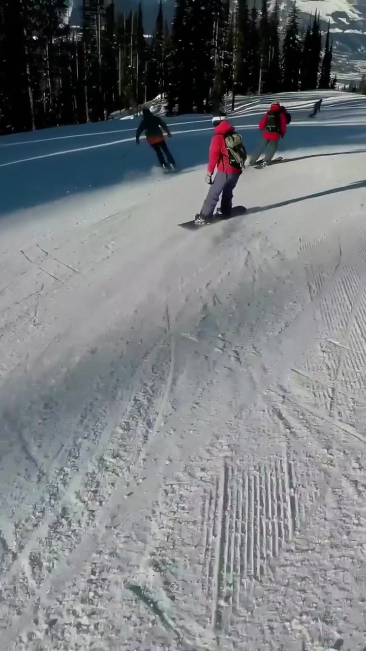 Video verticale: Snowboarder in giacca rossa che fa carving, rallentando sulla pista, con zaino, telecamera che si inclina.