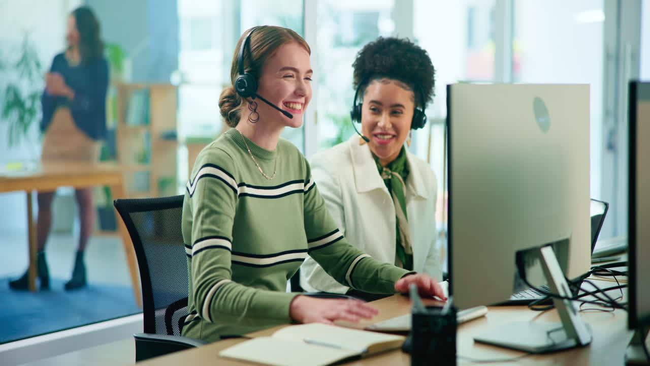 Two smiling women in headsets working at a computer in an office setting