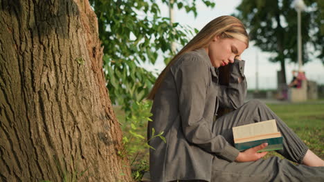 lady sitting outdoors under tree, reading book with hand under the book, legs stretched, head bent slightly with head resting on hand, greenery and blurred people walking in background