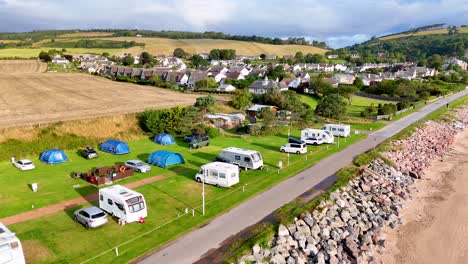 drone footage glides over a vibrant caravan and camping site near rosemarkie, scotland, revealing tents, motorhomes, and scenic countryside under bright daylight