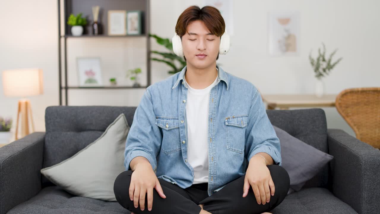 アジア人男性が白いヘッドフォンを着け、ソファで目を閉じて瞑想しています。