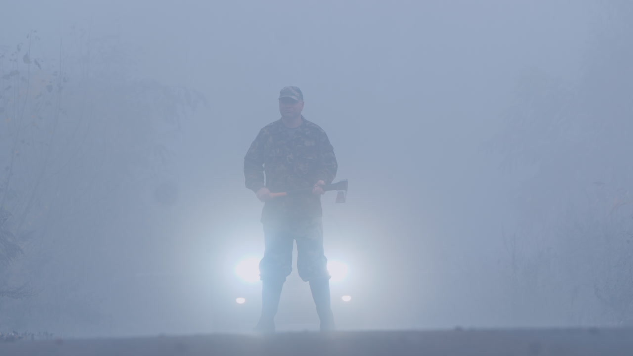 숲길에서 피 묻은 도끼를 든 살인자가 자동차 헤드라이트에 비추어 소름 끼치는 범죄 현장에 서 있다