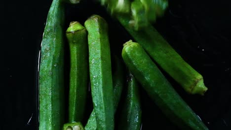 Dedos-Verdes-Cortados-En-Cámara-Lenta-Cayendo-Sobre-Okra-Entera-En-La-Superficie-Del-Agua-Aislada-En-Negro