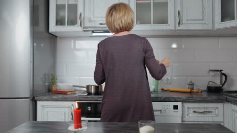 back view of house owner dancing in modern kitchen, wearing plaid clothing, surrounded by white cabinetry, countertop with rolling pin, kettle, and glassware, warm candlelight on island