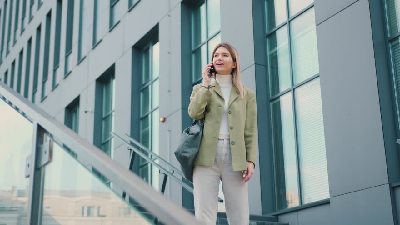 Pretty young smiling woman looking on watch while talking on smartphone during walking in urban city outdoors